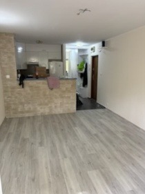 Jasne panele podłogowe w nowocześnie wykończonym wnętrzu z aneksem kuchennym i ścianą z dekoracyjnej cegły. Widok na przedpokój.