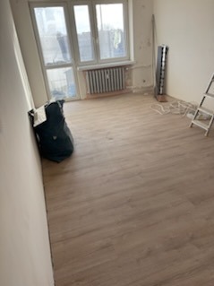 Jasne panele podłogowe w pokoju przed wykończeniem ścian. Widoczne ślady po gładzi, okno z grzejnikiem, drabina i worek na śmieci. Ujęcie z dołu.