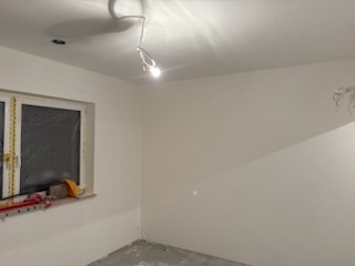 Białe, świeżo pomalowane ściany w pokoju z oknem. Widoczne ślady po remoncie na podłodze i niedokończone wykończenie przy oknie. Oświetlenie sufitowe.