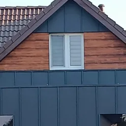 Fragment elewacji domu z ciemnoszarą blachą trapezową, drewnianą deską poddasza i oknem dwuskrzydłowym, dach pokryty brązową dachówką ceramiczną.