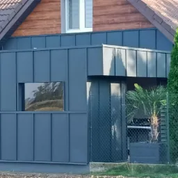 Nowoczesny dom z ciemnoszarą elewacją z paneli na rąbek stojący, brązowym dachem z dachówki i drewnianym wykończeniem pod dachem, widok zza ogrodzenia.