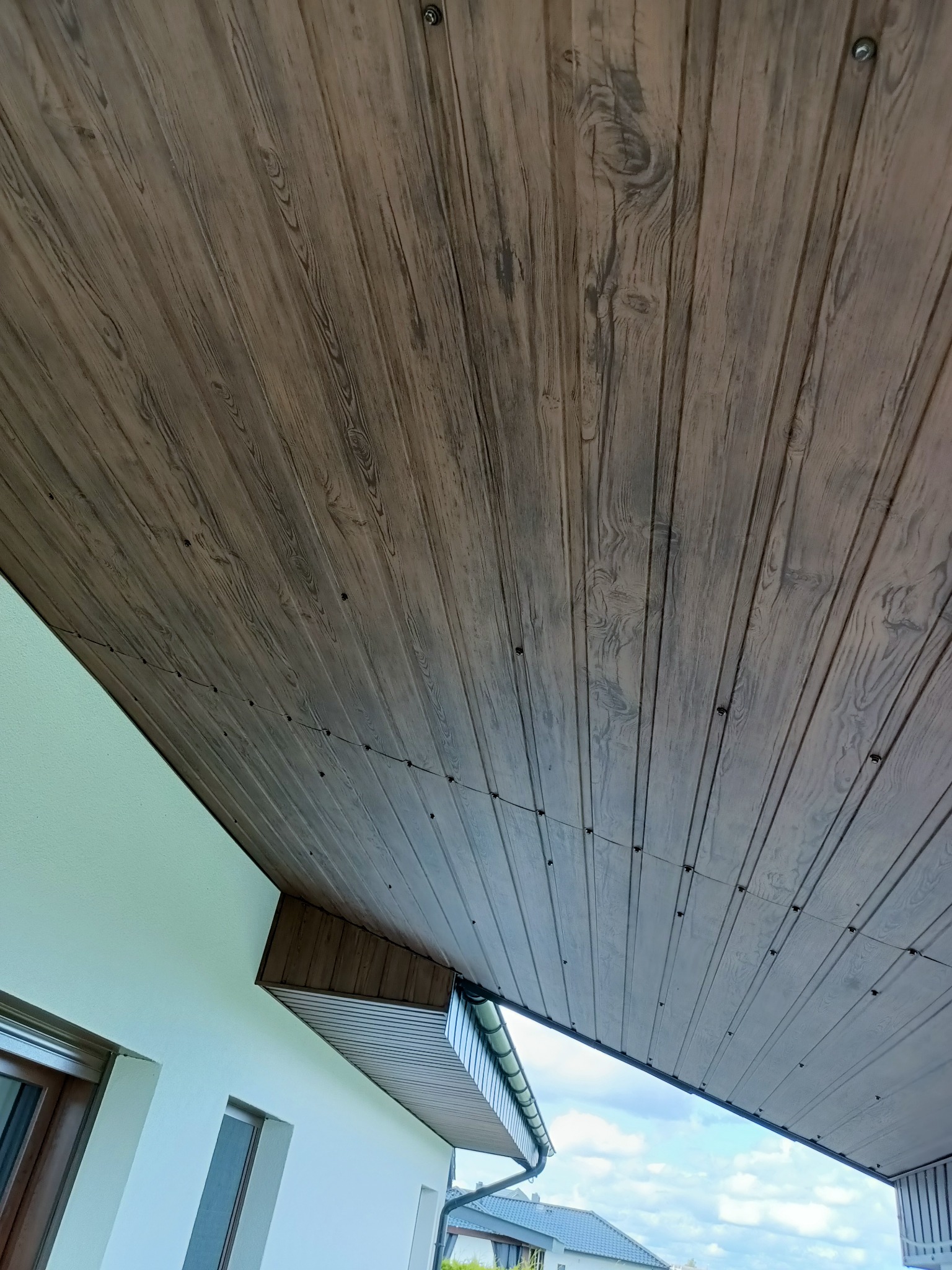 Podbitka dachowa z paneli drewnopodobnych, mocowana śrubami, widok od dołu, fragment ściany budynku w tle.