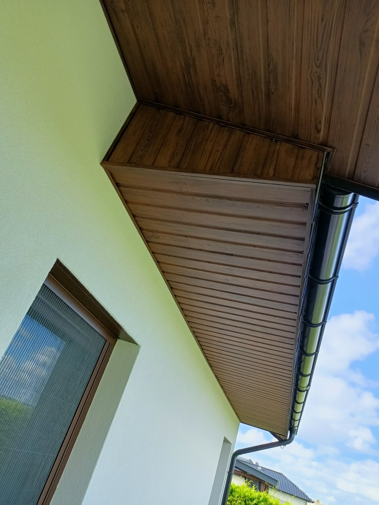 Podbitka dachowa z paneli imitujących drewno, połączona z ciemną rynną, widok z dołu na tle błękitnego nieba.