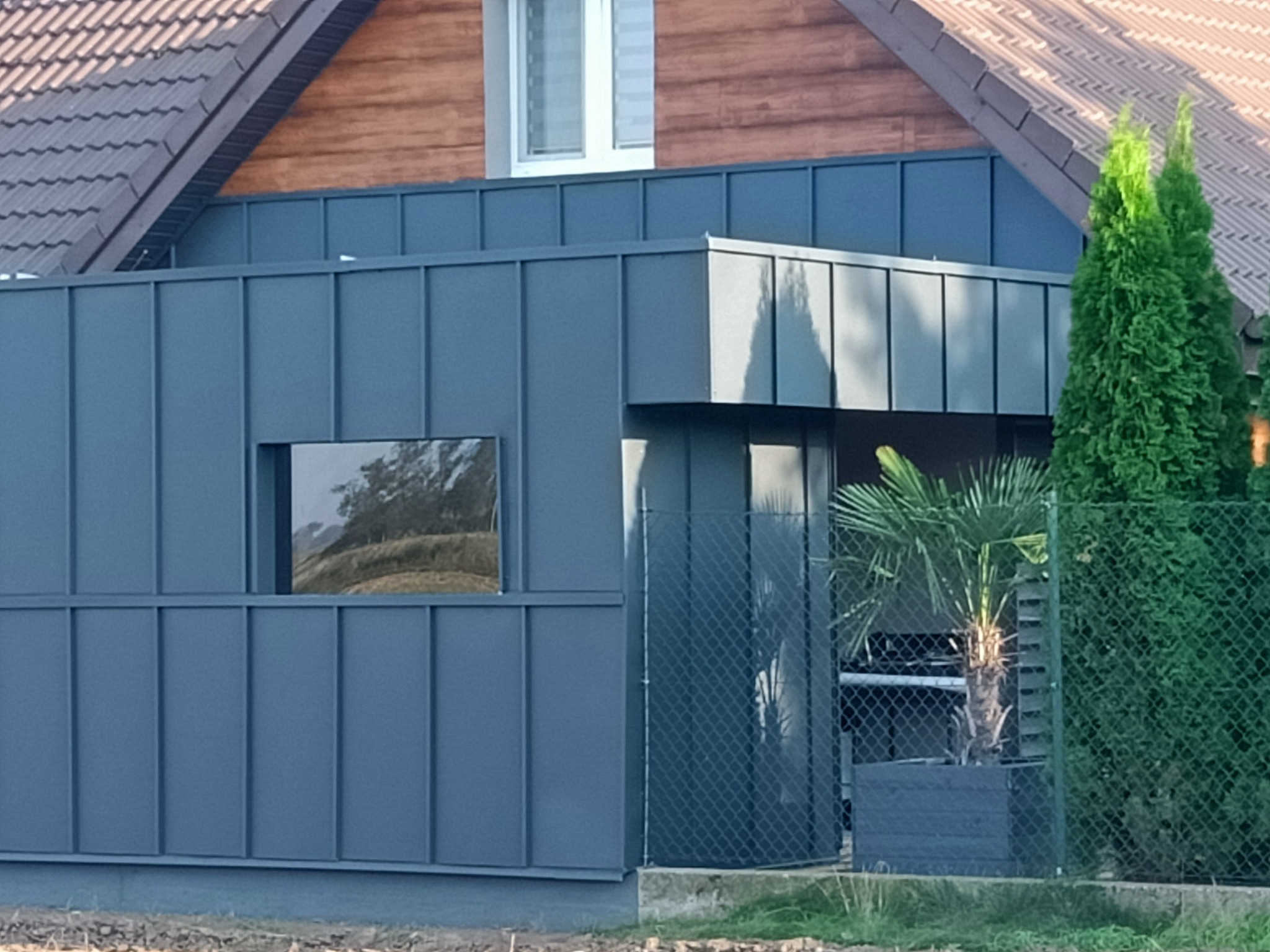 Nowoczesny dom z ciemnoszarą elewacją z paneli na rąbek stojący, brązowym dachem z dachówki i drewnianym wykończeniem pod dachem, widok zza ogrodzenia.