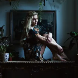 Kobieta z blond włosami w kwiecistej sukience siedzi na zdobionym stole w otoczeniu roślin doniczkowych i obrazu w ramie, spoglądając w bok.