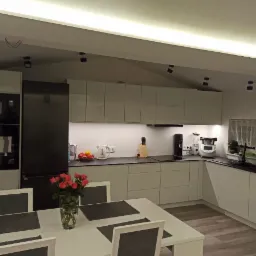 Jasna kuchnia z zabudową pod skosem, czarny sprzęt AGD, bukiet róż na stole, oświetlenie punktowe i listwowe.