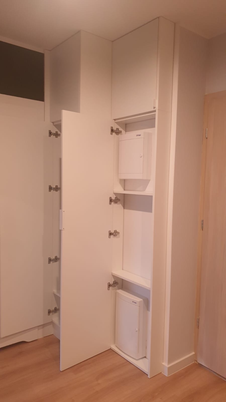 Biała szafa wnękowa z otwartymi drzwiami, ukazująca zamontowane skrzynki elektryczne na półkach, widoczne zawiasy i drewnopodobna podłoga.