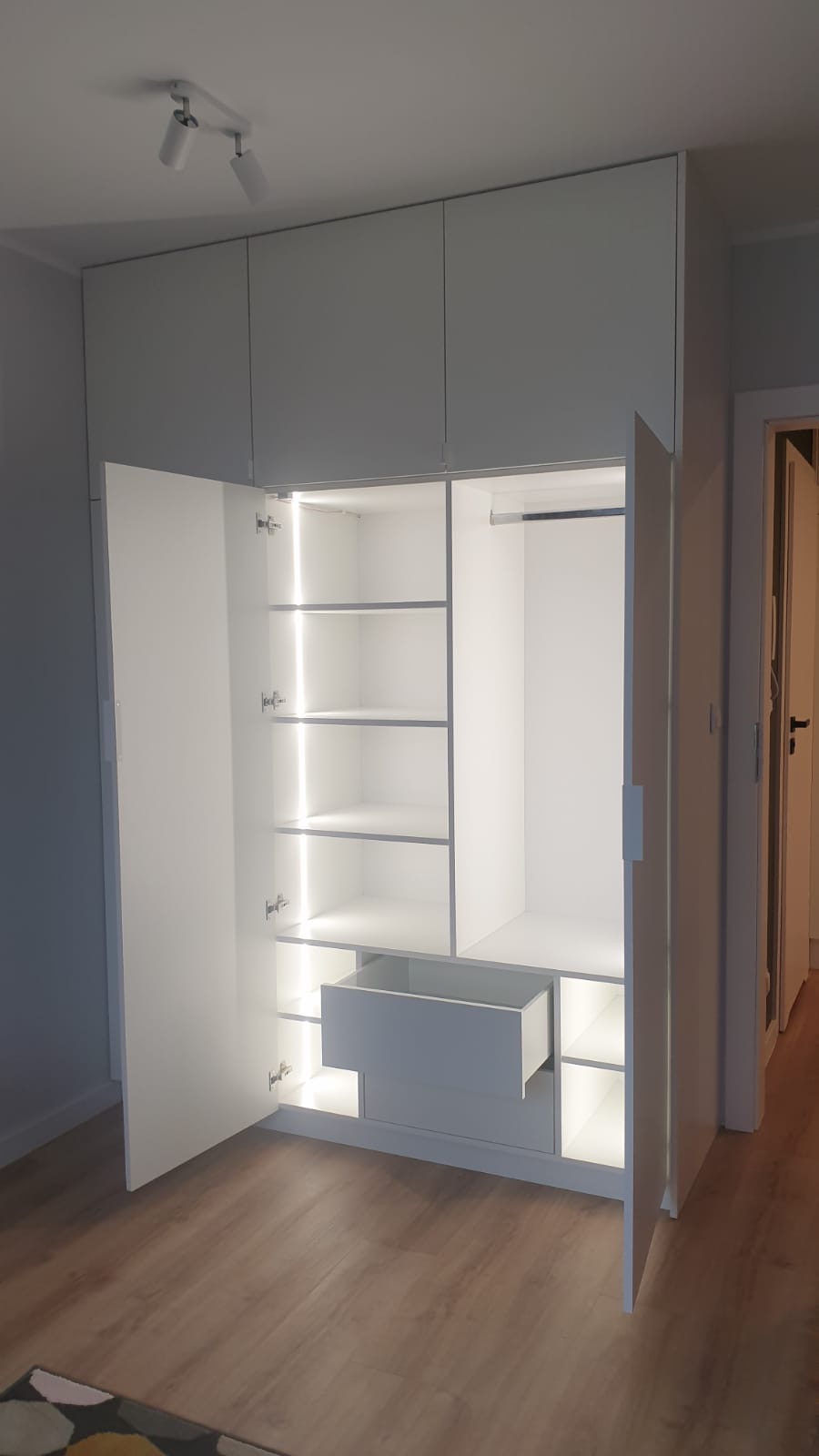 Biała szafa wnękowa z otwartymi drzwiami, podświetlane półki i wysuwana szuflada, widok na drewnianą podłogę i fragment dywanu.