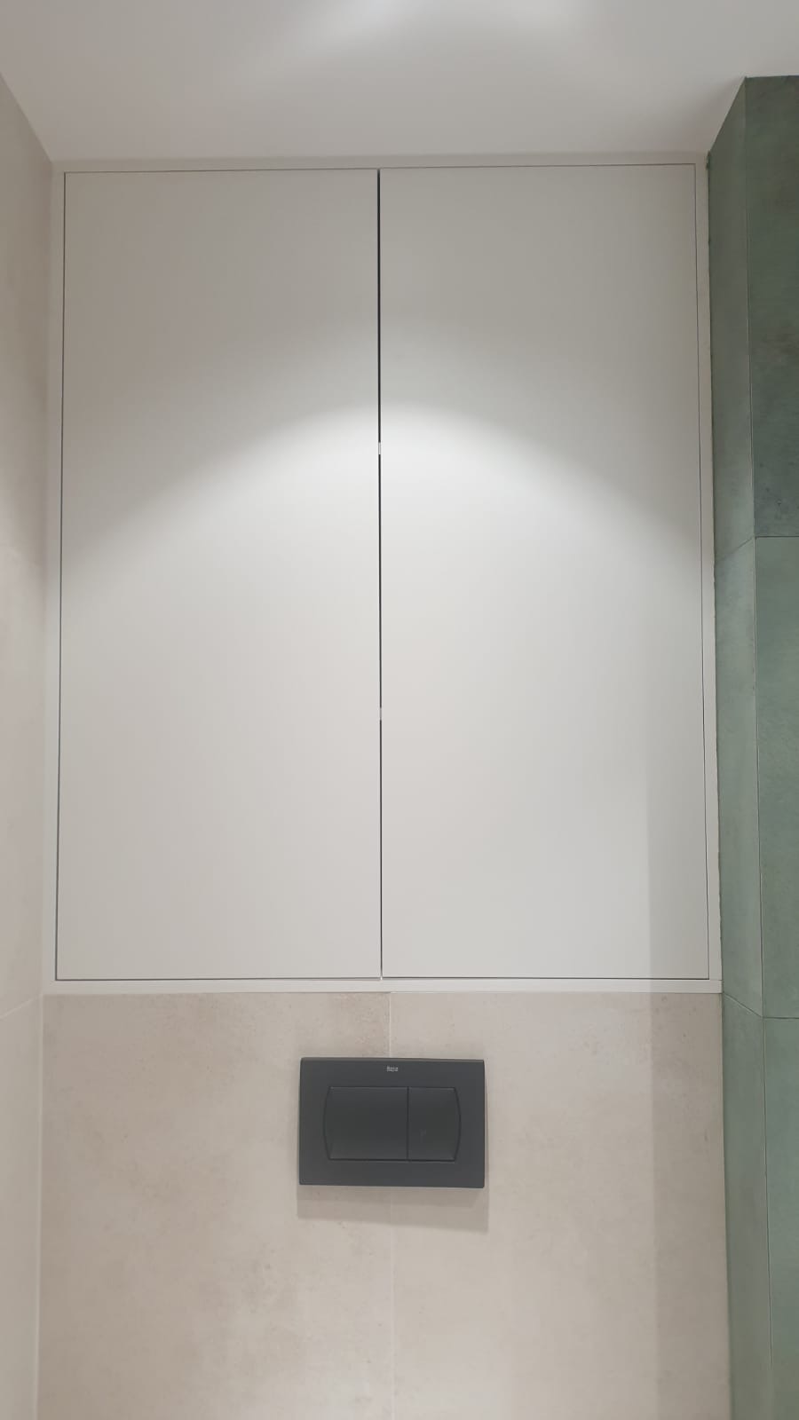 Biała szafka w zabudowie nad toaletą z przyciskiem spłukującym Roca, na tle beżowych i zielonych płytek.