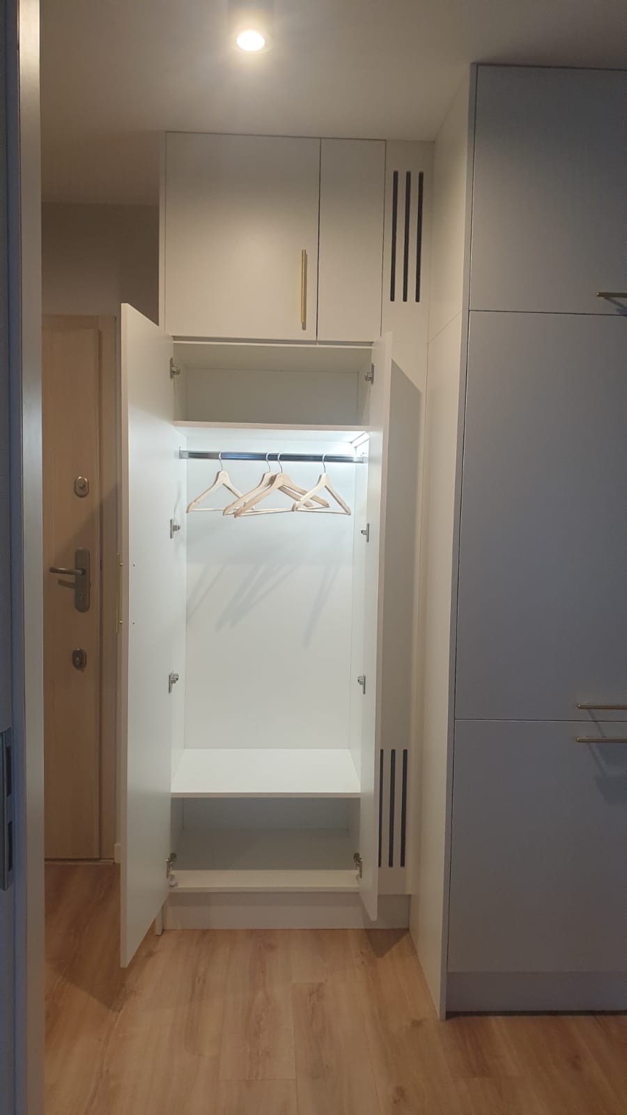 Wnętrze białej szafy wnękowej z otwartymi drzwiami, widoczne trzy drewniane wieszaki, metalowy drążek i półka, obok szara szafa z podłużnymi uchwytami.