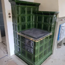 Zielony piec kaflowy z żeliwną płytą grzewczą i złotym relingiem, widoczny w trakcie montażu, z odsłoniętymi przewodami elektrycznymi w ścianie.