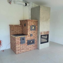 Manufaktura Ognia s.c. - Nowo wybudowany piec kaflowy z cegły z żeliwnymi drzwiczkami i kominek z szarego betonu w surowym wnętrzu z widocznymi elementami instalacji na ścianie.