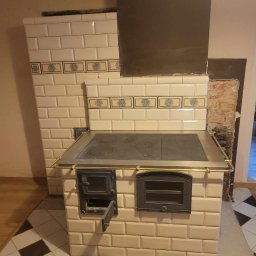 Manufaktura Ognia s.c. - Tradycyjny piec kaflowy z metalową płytą grzewczą i otwartymi drzwiczkami paleniska, obudowany beżowymi kaflami z dekoracyjnym pasem kwiatowym, z widocznym fragmentem komina i surowej ściany w tle.