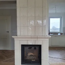 Biały, kaflowy kominek z czarnym wkładem, dekoracyjne płytki, widoczne ślady prac wykończeniowych na podłodze.