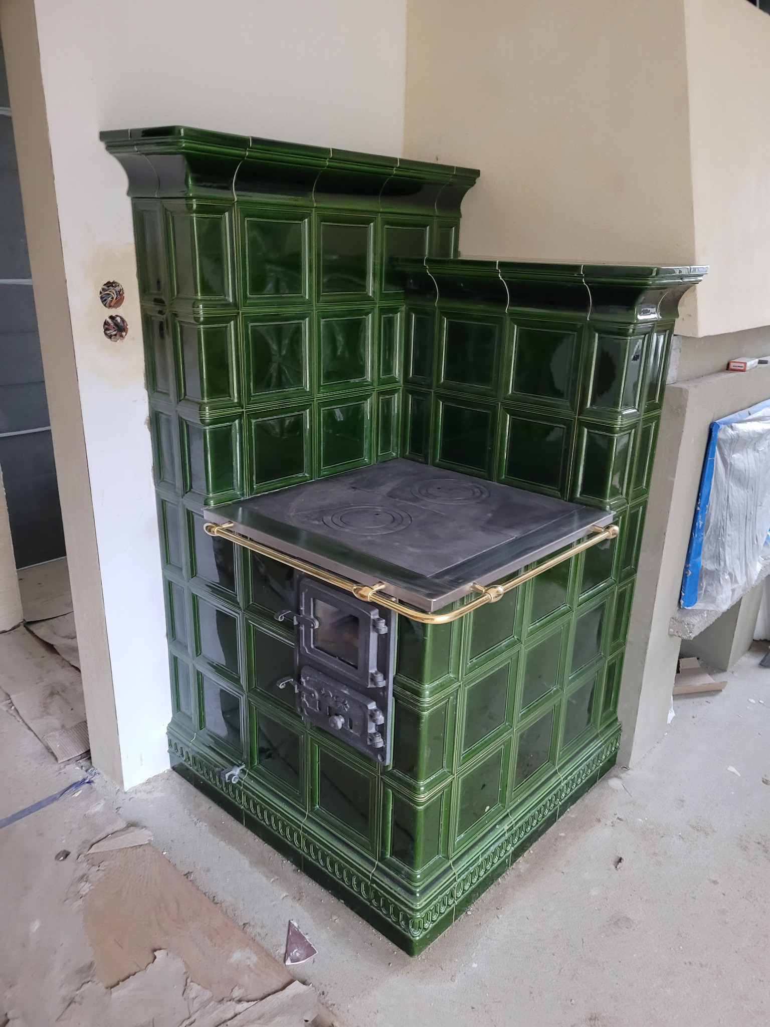 Zielony piec kaflowy z żeliwną płytą grzewczą i złotym relingiem, widoczny w trakcie montażu, z odsłoniętymi przewodami elektrycznymi w ścianie.