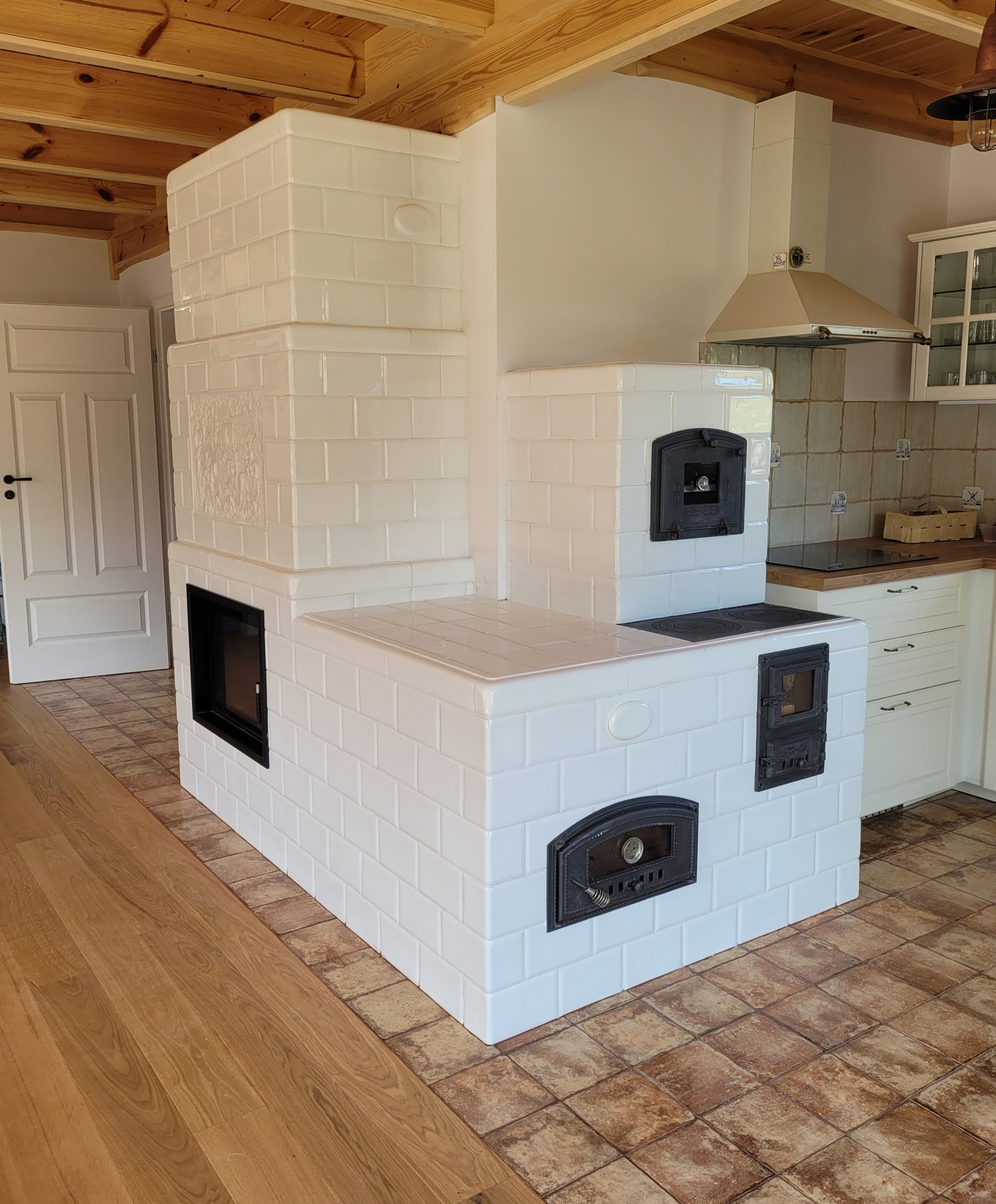 Biały piec kaflowy z czarnymi drzwiczkami w kuchni, widoczne drewniane belki stropowe i fragment zabudowy kuchennej