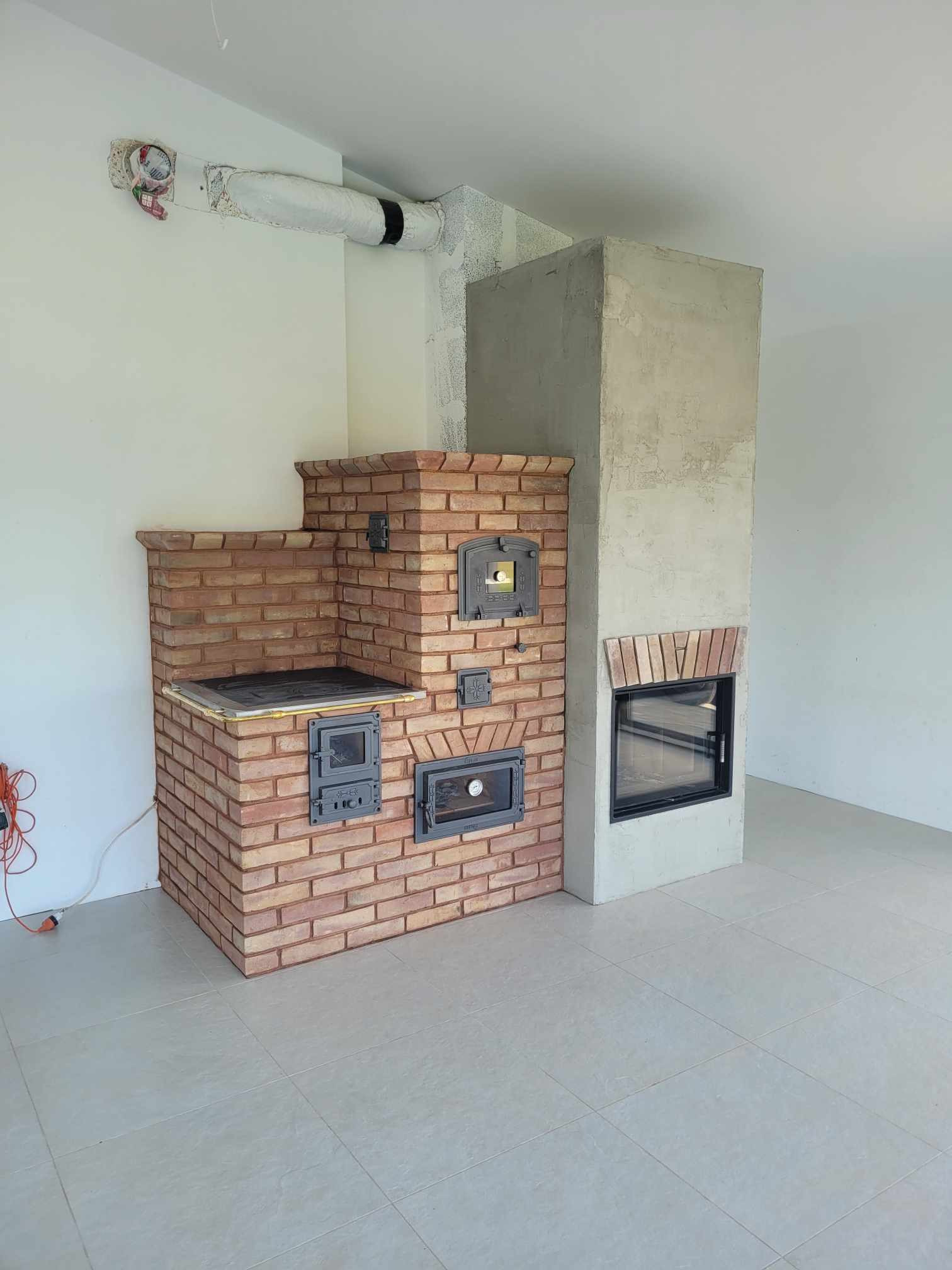 Nowo wybudowany piec kaflowy z cegły z żeliwnymi drzwiczkami i kominek z szarego betonu w surowym wnętrzu z widocznymi elementami instalacji na ścianie.