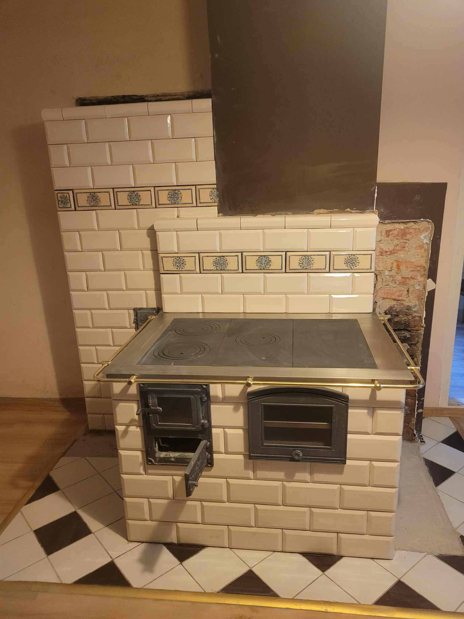 Tradycyjny piec kaflowy z metalową płytą grzewczą i otwartymi drzwiczkami paleniska, obudowany beżowymi kaflami z dekoracyjnym pasem kwiatowym, z widocznym fragmentem komina i surowej ściany w tle.