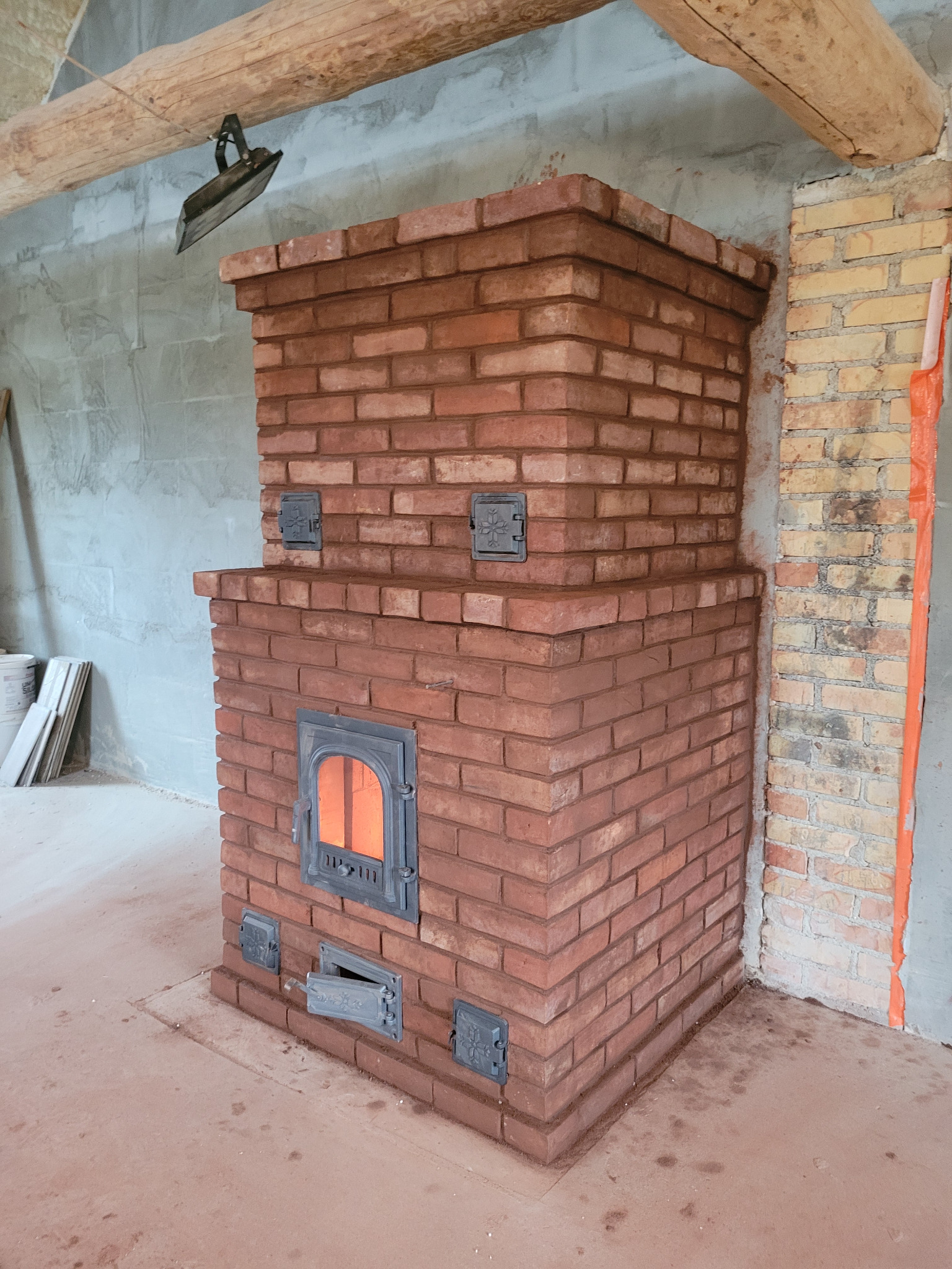 Nowy piec z czerwonej cegły w trakcie budowy, z widocznym paleniskiem i metalowymi drzwiczkami popielnika, na tle surowych ścian i drewnianej belki stropowej.