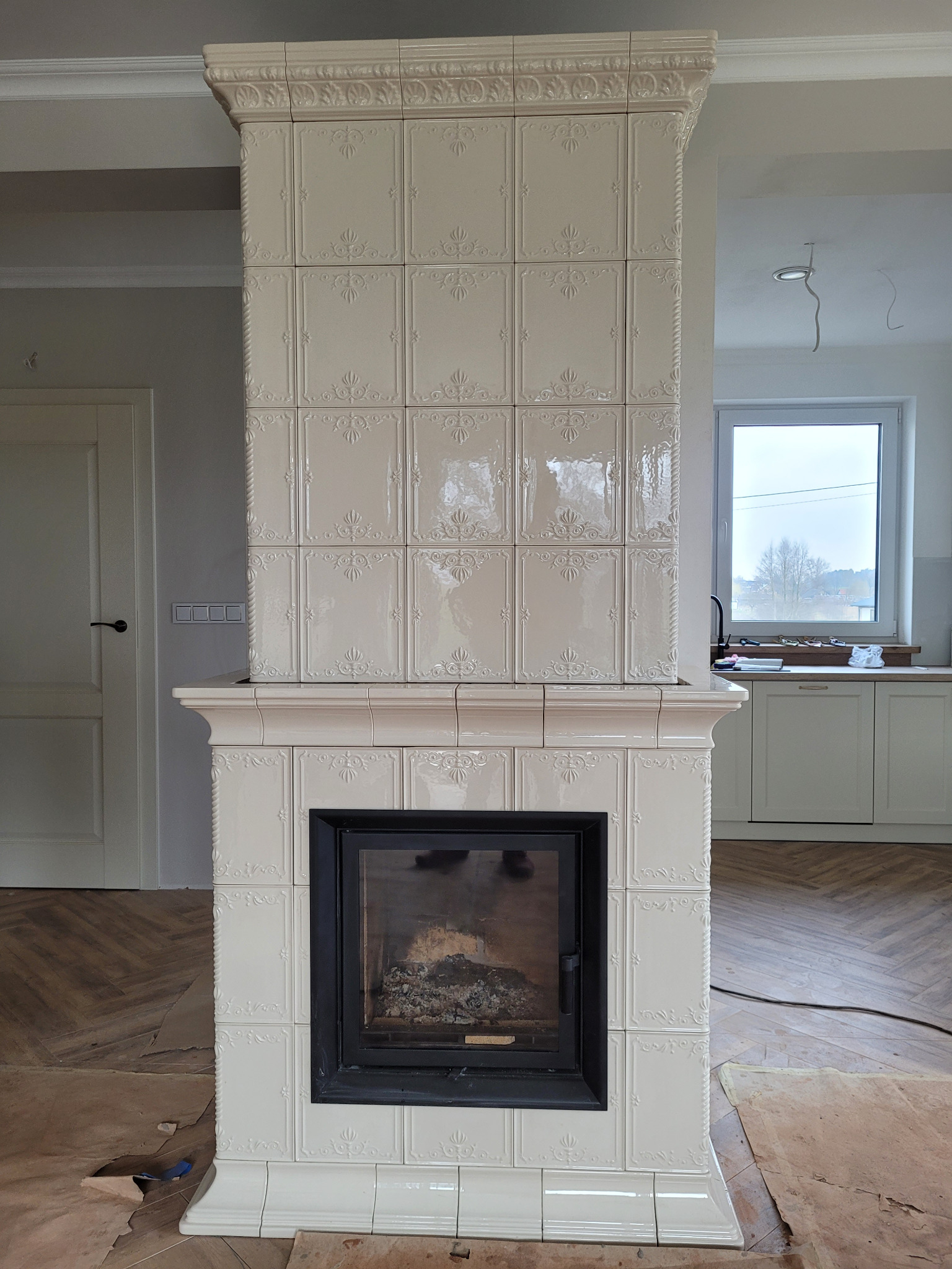 Biały, kaflowy kominek z czarnym wkładem, dekoracyjne płytki, widoczne ślady prac wykończeniowych na podłodze.