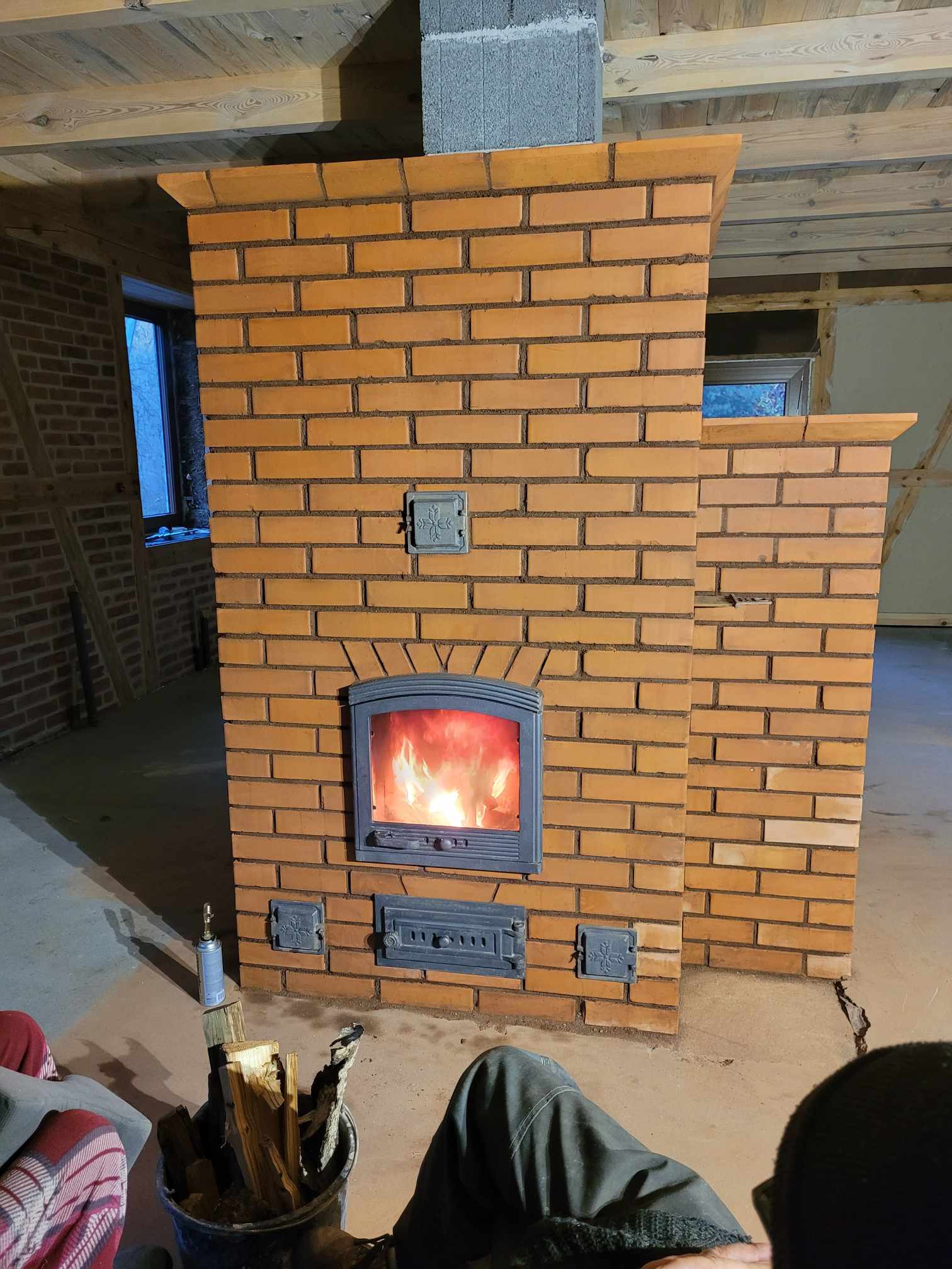 Nowo wybudowany, ceglany piec z żeliwnymi drzwiczkami, widoczne płomienie w palenisku, obok wiadro z drewnem i butla gazowa.