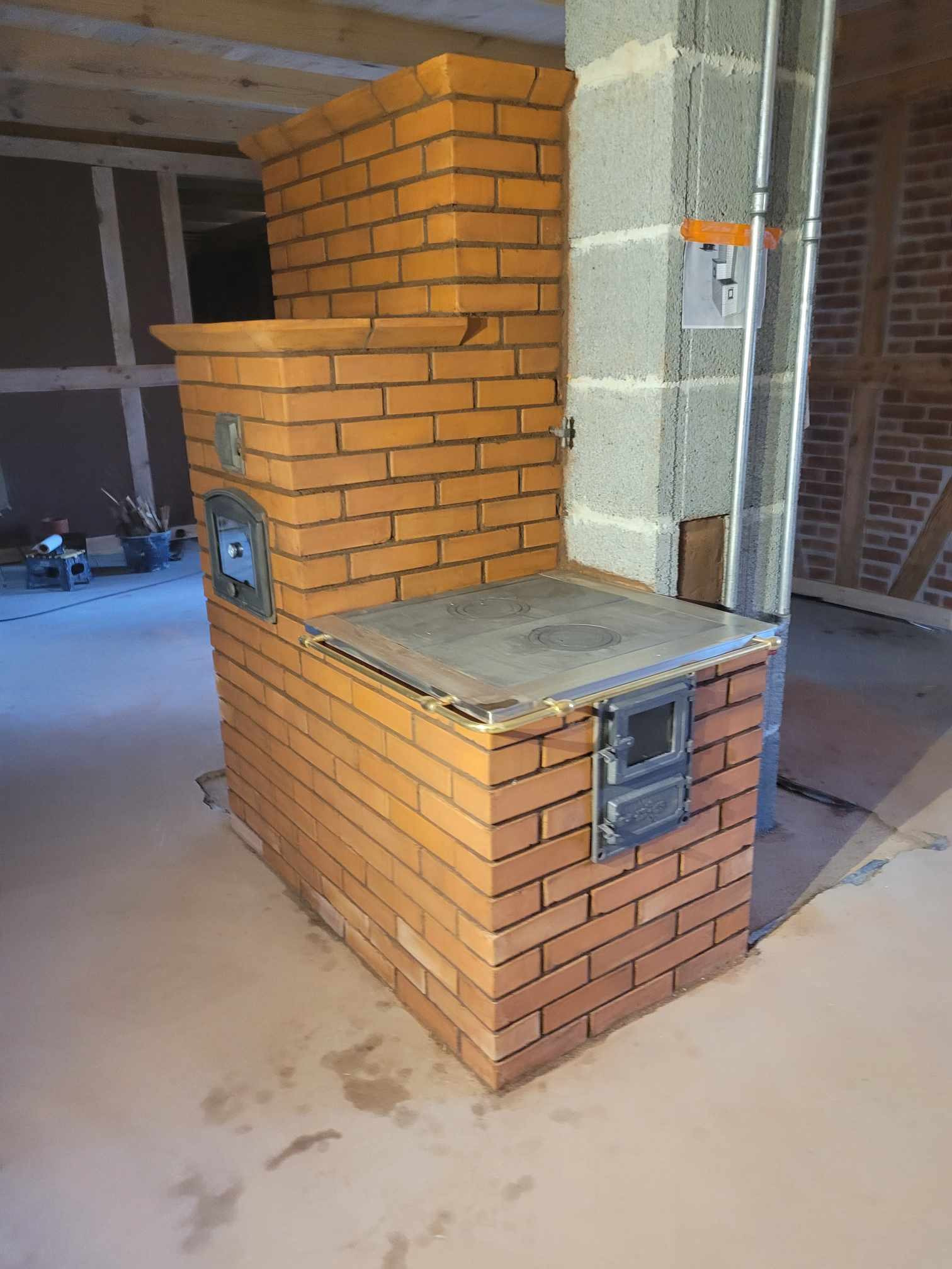 Nowo wybudowany, ceglany piec kuchenny z metalową płytą grzewczą i drzwiczkami, widoczny w surowym wnętrzu z betonową podłogą i elementami konstrukcyjnymi w tle.