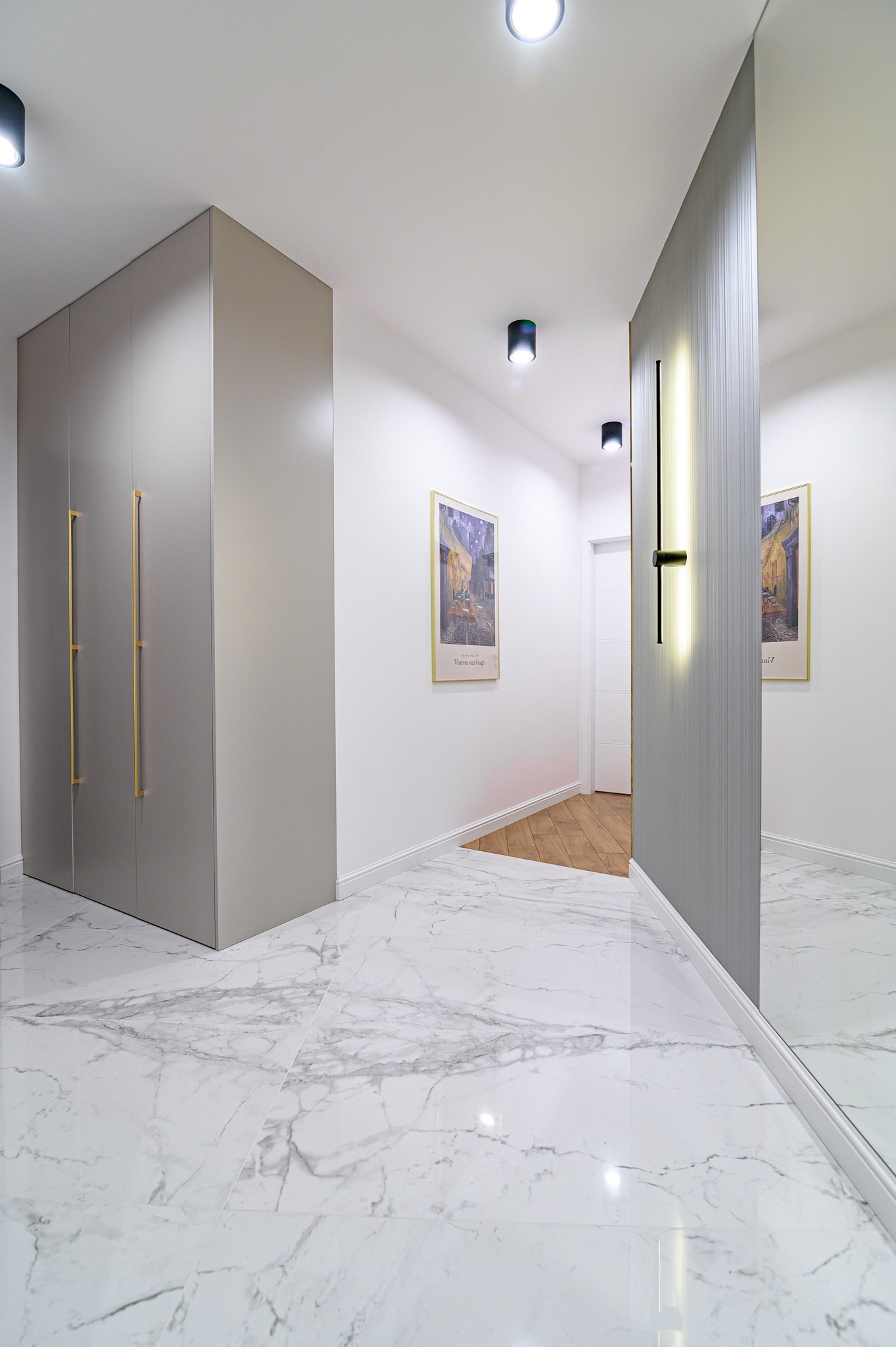 Nowoczesny przedpokój z szarą szafą w zabudowie, marmurową podłogą i lustrem odbijającym obraz Van Gogha.