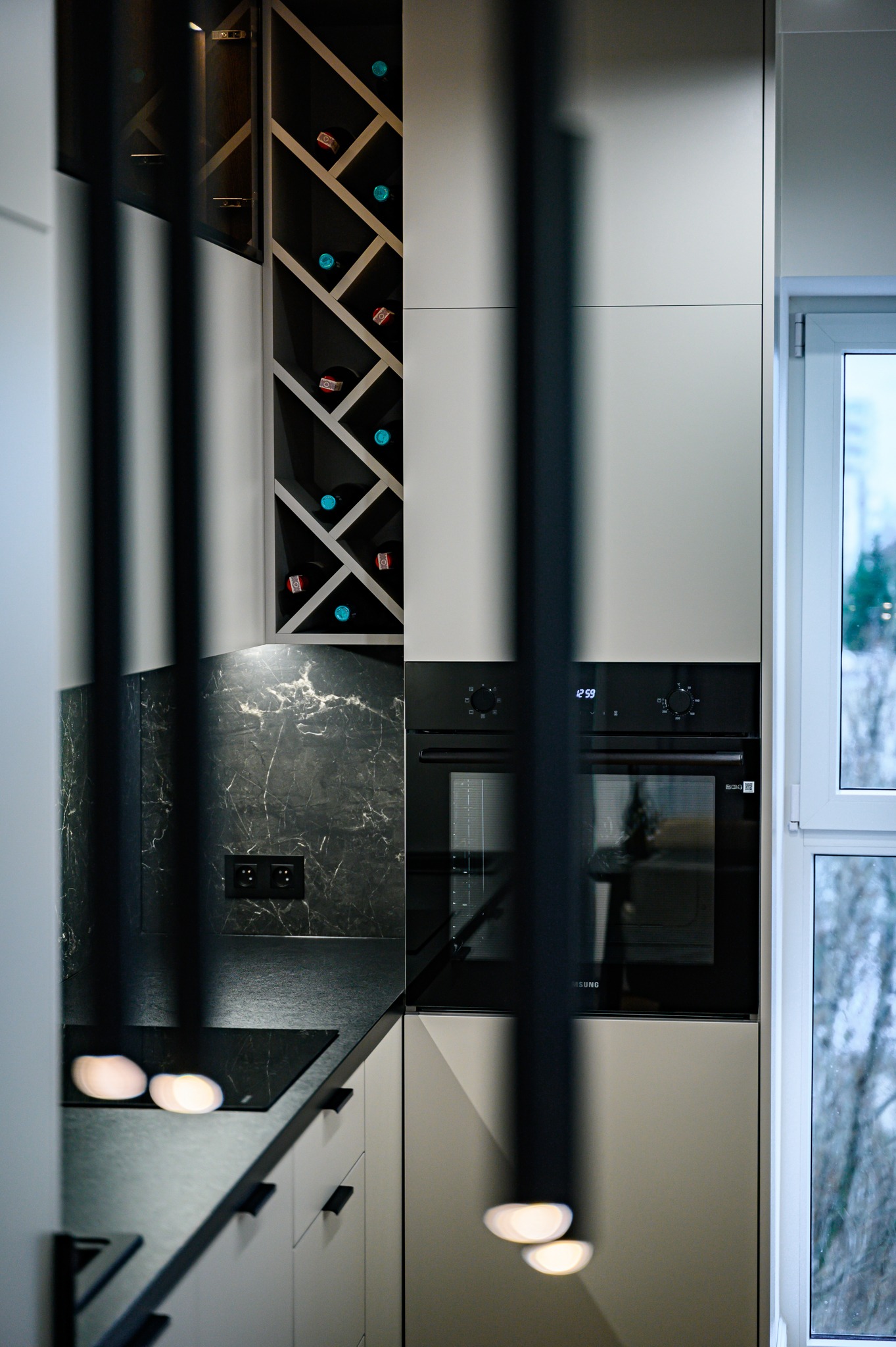 Nowoczesna kuchnia z białymi frontami, czarnym blatem imitującym marmur, zabudowanym piekarnikiem i regałem na wino, widziana przez dekoracyjne, pionowe elementy oświetleniowe.