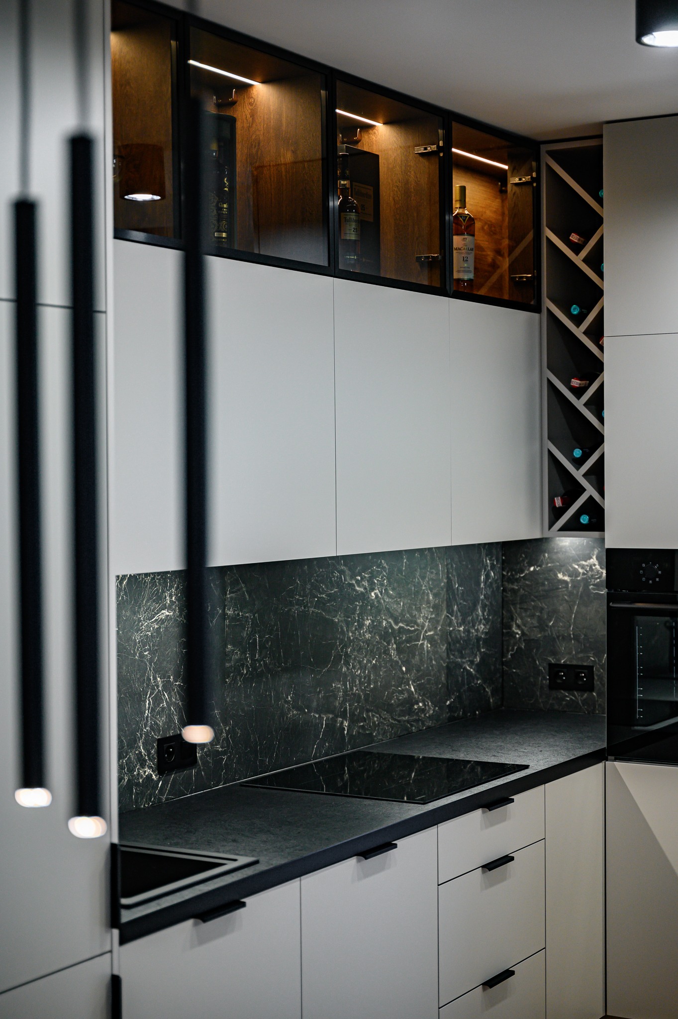 Nowoczesna kuchnia z białymi szafkami, czarnym blatem imitującym marmur i podświetlanymi witrynami z ekspozycją alkoholi.