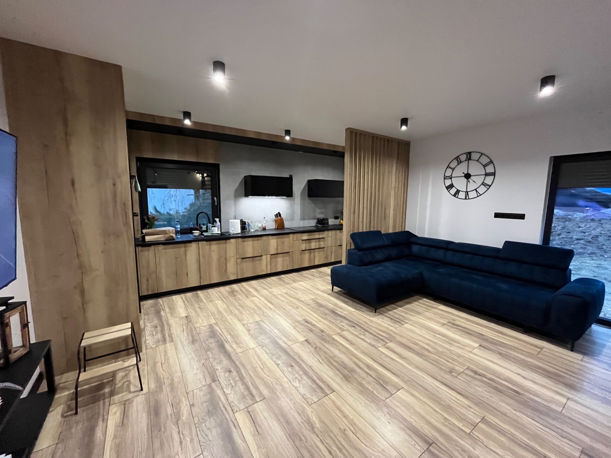 Wnętrze salonu z otwartą kuchnią: drewniane szafki kuchenne z czarnymi akcentami, narożna sofa w odcieniach granatu, drewnopodobna podłoga i dekoracyjny zegar ścienny.