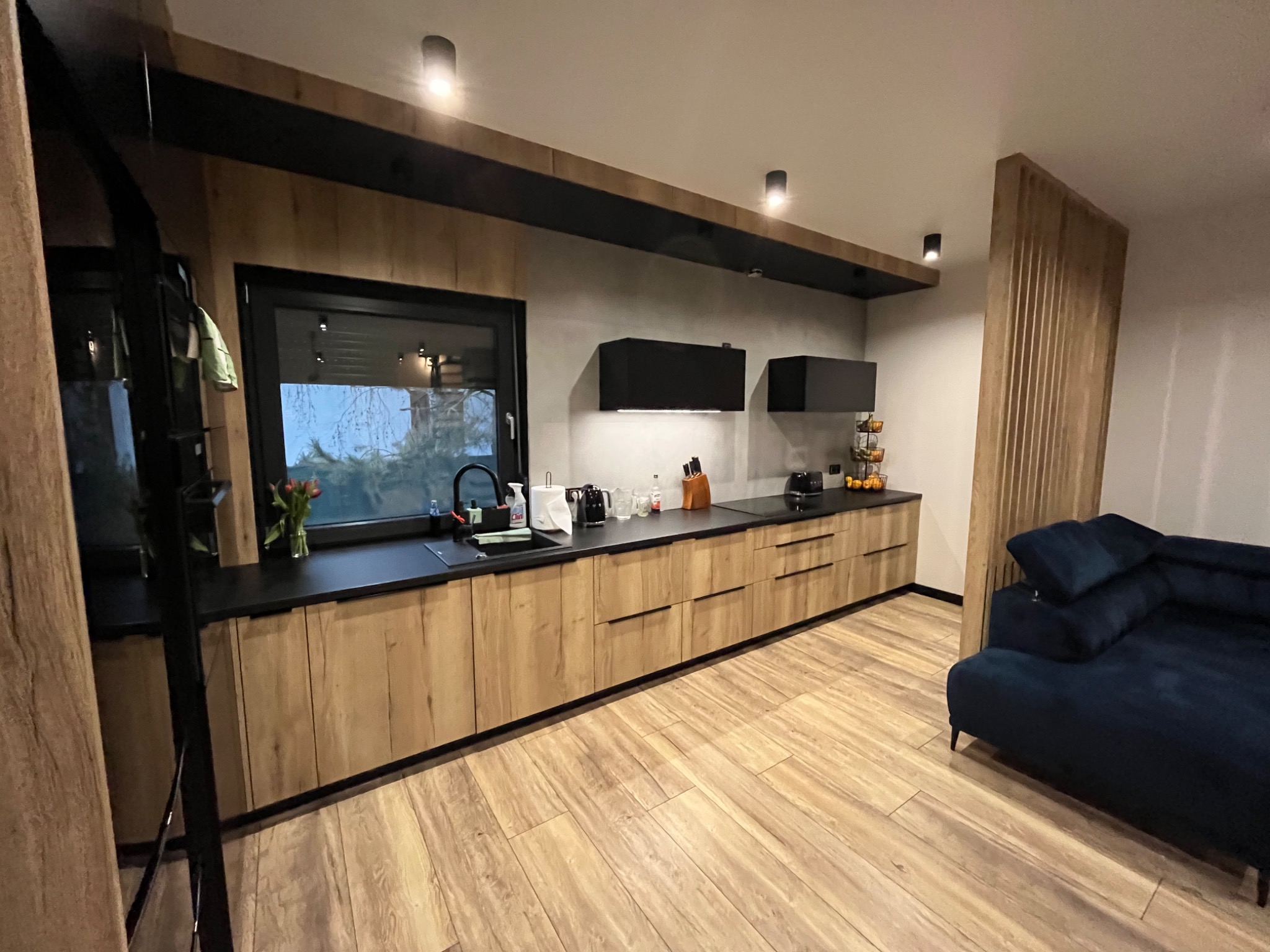 Nowoczesna kuchnia z drewnianymi frontami szafek, czarnym blatem i elementami dekoracyjnymi z drewna, oświetlona punktowymi lampami sufitowymi, z widoczną częścią salonu z granatową sofą.