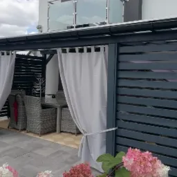 Nowoczesna, ciemnoszara konstrukcja z poziomych listew, z zasłonami po bokach, tworząca zaciszną strefę relaksu z rattanowymi meblami ogrodowymi i szklanym stolikiem, otoczona kwitnącymi hortensjami.