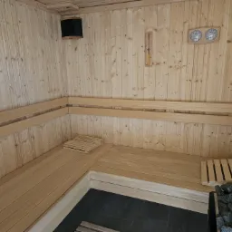 Wnętrze sauny wykończone jasnym drewnem, z drewnianymi ławkami ułożonymi w kształt litery L, czarną podłogą z płytek, termometrem i higrometrem na ścianie oraz kamieniami w piecu.