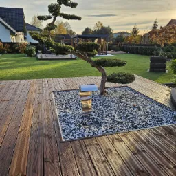 Drewniany taras z wbudowanym klombem wypełnionym białymi kamieniami i ozdobnym drzewkiem bonsai, w tle widoczna altana i zadbany trawnik.