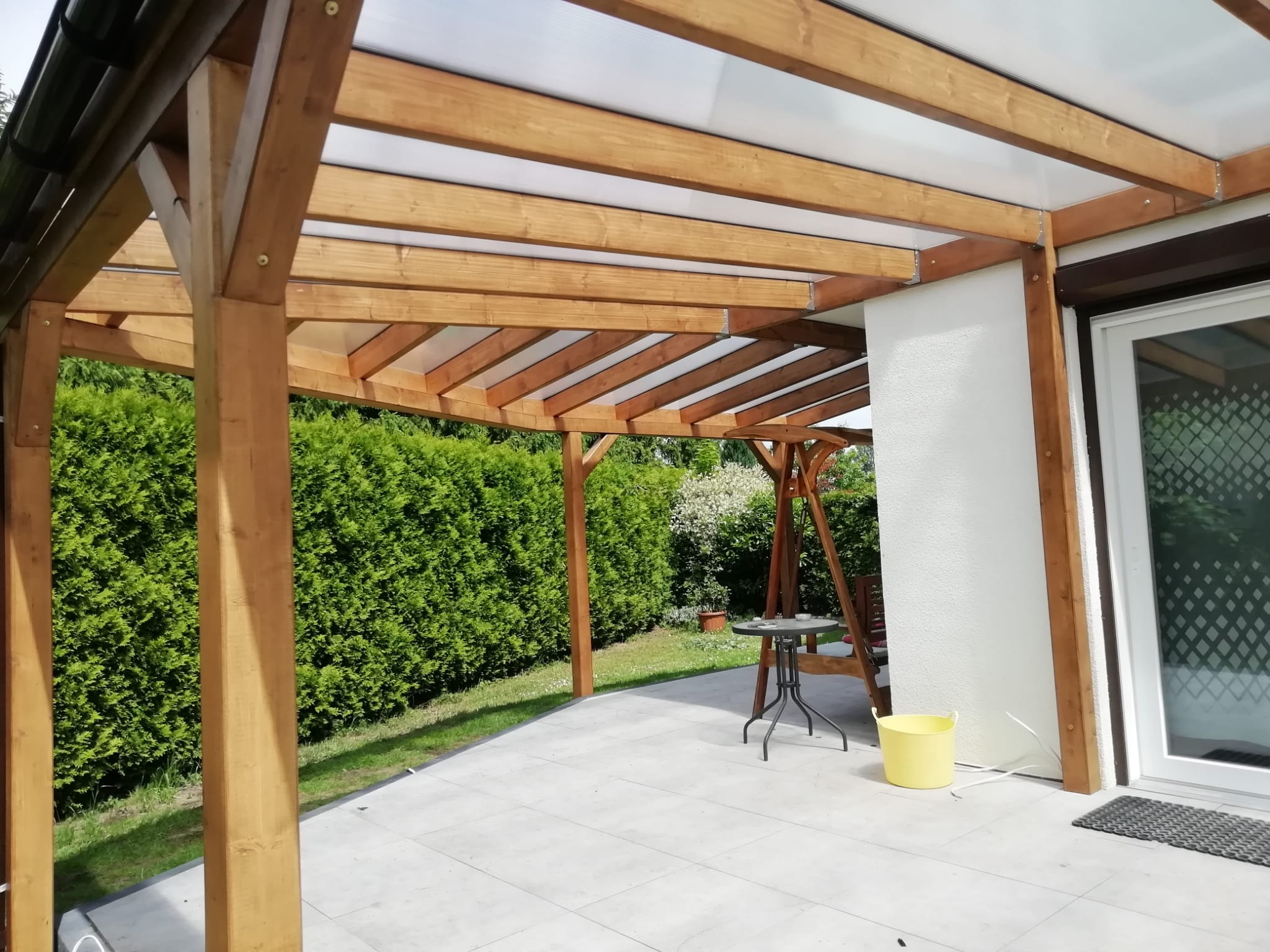 Drewniana konstrukcja z przezroczystym zadaszeniem tworząca zacienione miejsce do wypoczynku na tarasie z jasnymi płytkami, obok domu z białymi ścianami i zielonym żywopłotem w tle. Widoczny stolik...