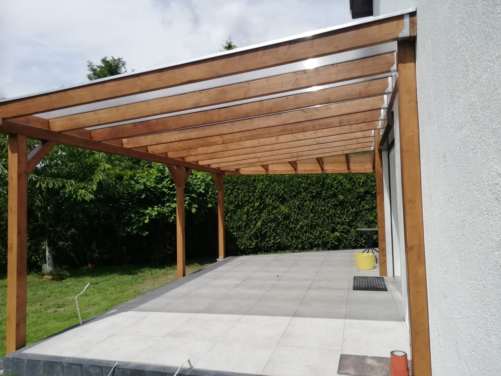 Drewniana konstrukcja z przezroczystym zadaszeniem przylegająca do białego budynku, z widocznym fragmentem tarasu wyłożonego szarą kostką.