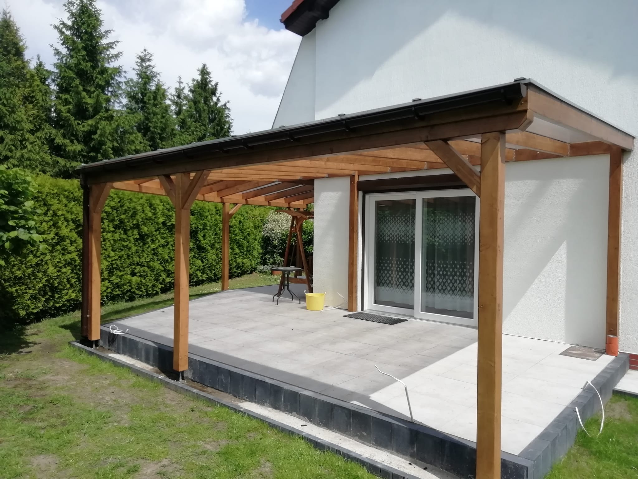 Drewniana konstrukcja z zadaszeniem przylegająca do białego domu z przesuwnymi drzwiami tarasowymi, widoczny fragment tarasu z szarych płyt, zielony trawnik i drzewa w tle.