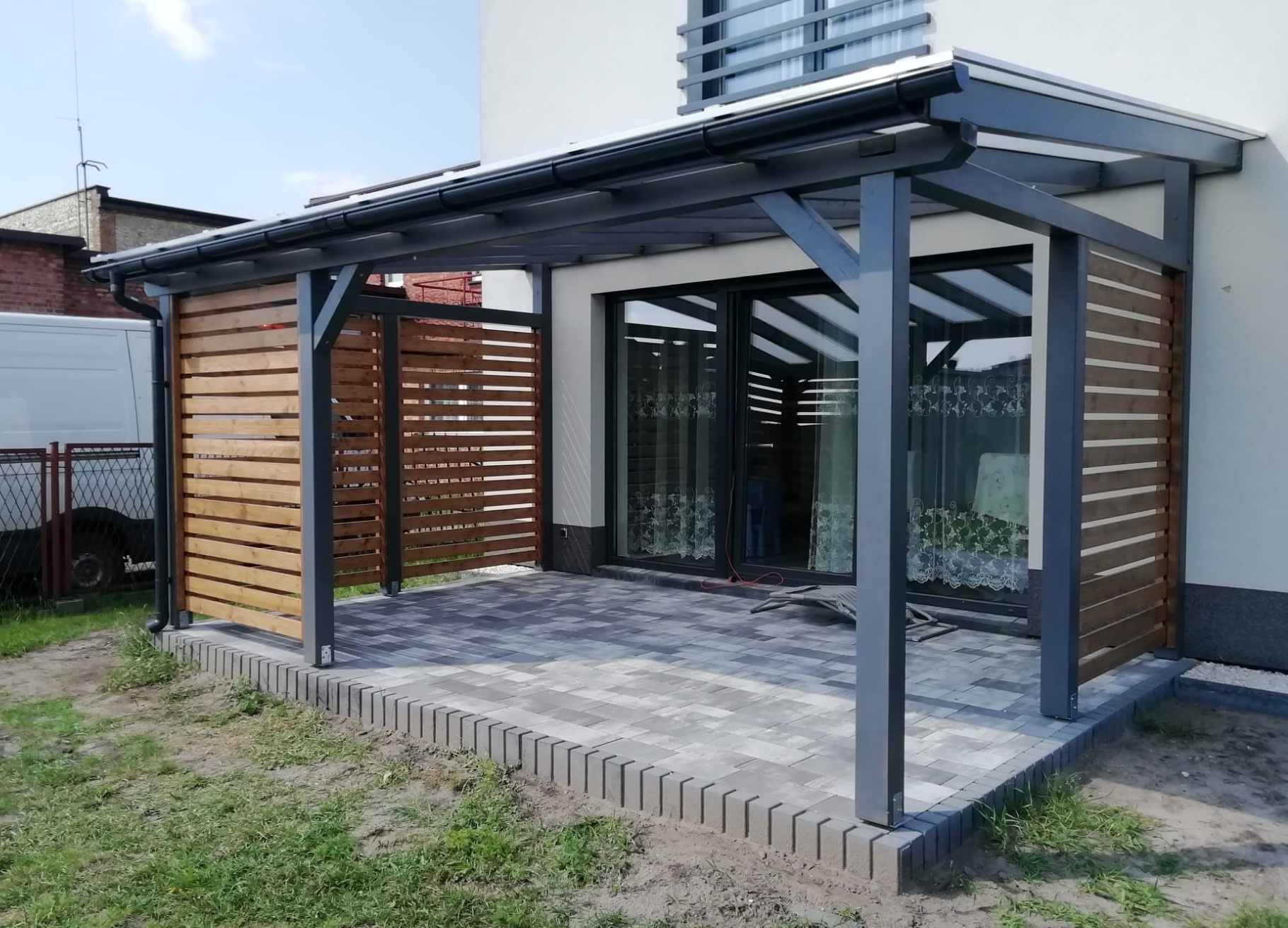 Zadaszona konstrukcja z drewnianymi panelami bocznymi i szarą ramą, przylegająca do budynku z przesuwnymi drzwiami, wyłożona szarą kostką brukową.