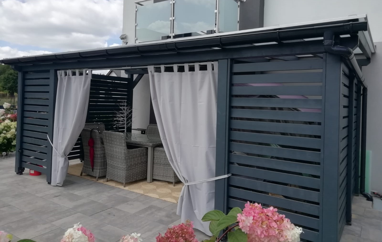 Nowoczesna, ciemnoszara konstrukcja z poziomych listew, z zasłonami po bokach, tworząca zaciszną strefę relaksu z rattanowymi meblami ogrodowymi i szklanym stolikiem, otoczona kwitnącymi hortensjami.