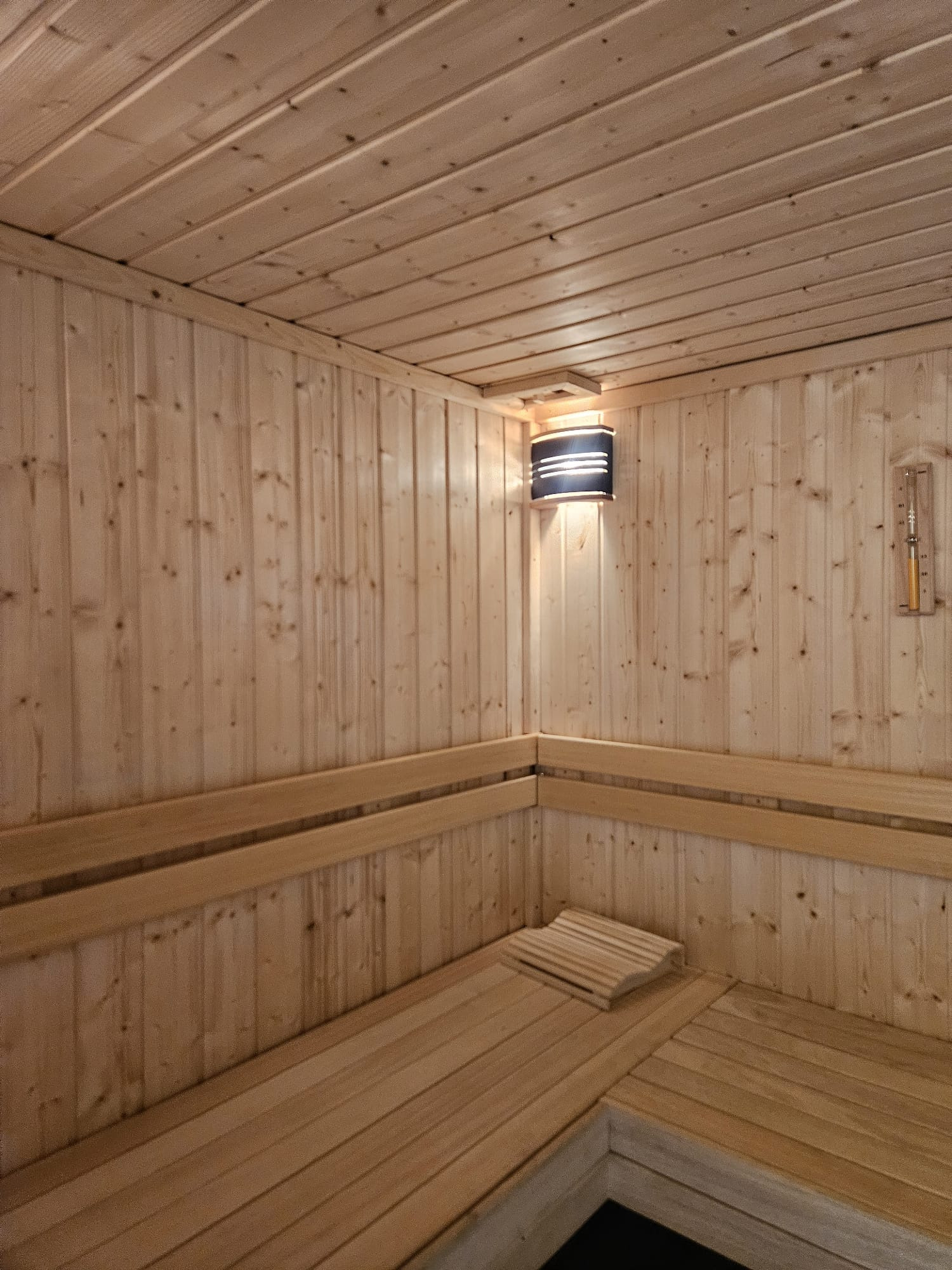 Wnętrze sauny z jasnego drewna, widoczne ławy, oświetlenie ścienne i termometr. Drewniana poduszka na siedzisku.