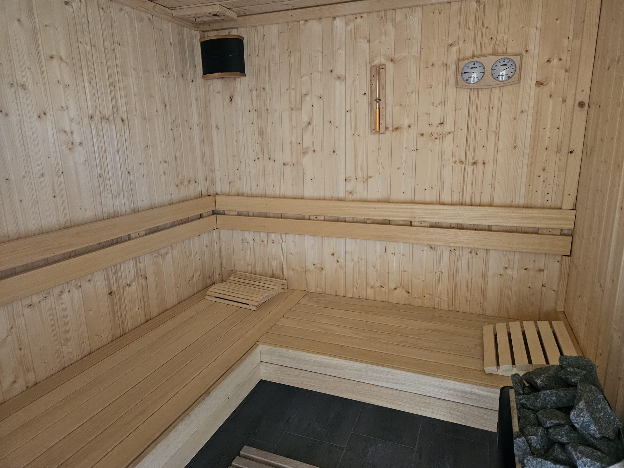 Wnętrze sauny wykończone jasnym drewnem, z drewnianymi ławkami ułożonymi w kształt litery L, czarną podłogą z płytek, termometrem i higrometrem na ścianie oraz kamieniami w piecu.