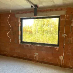 Surowy stan wnętrza z widocznymi przewodami elektrycznymi wystającymi ze ścian i sufitu, okno z widokiem na drzewa, ściany z czerwonej cegły dziurawki.