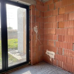 Shine Adam Juraszczyk - Surowy stan wnętrza: ściana z czerwonej cegły z przygotowanymi punktami elektrycznymi, widoczne przewody, okno z czarną ramą, wylewka betonowa na podłodze.