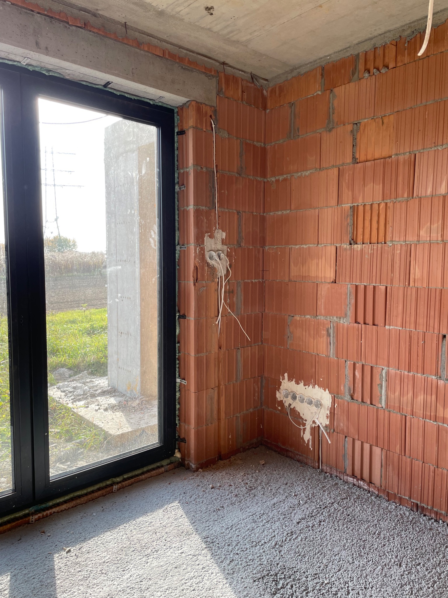 Surowy stan wnętrza: ściana z czerwonej cegły z przygotowanymi punktami elektrycznymi, widoczne przewody, okno z czarną ramą, wylewka betonowa na podłodze.