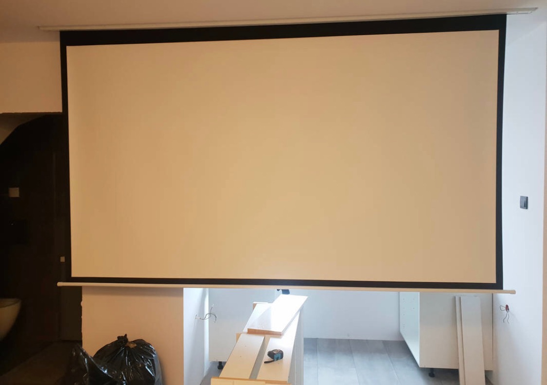 Zamontowany ekran projekcyjny w pomieszczeniu w trakcie prac wykończeniowych, widoczne elementy instalacji elektrycznej i fragmenty mebli.