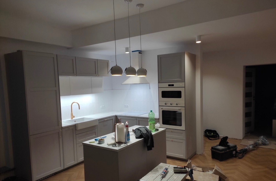 Szara zabudowa kuchenna z wyspą, oświetleniem punktowym i trzema lampami wiszącymi, widoczne narzędzia i materiały wykończeniowe sugerują etap prac wykończeniowych.