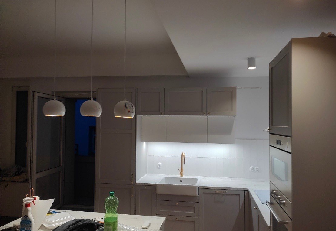 Nowoczesna kuchnia w trakcie wykańczania: szare szafki, białe płytki nad blatem, złota bateria przy zlewie, trzy wiszące białe lampy kuliste, widoczne materiały budowlane na blacie.