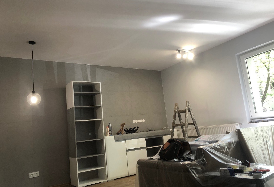 Wnętrze pokoju w trakcie remontu, widoczna drabina, meble zabezpieczone folią, wisząca lampa z okrągłym kloszem i sufitowe oświetlenie punktowe.