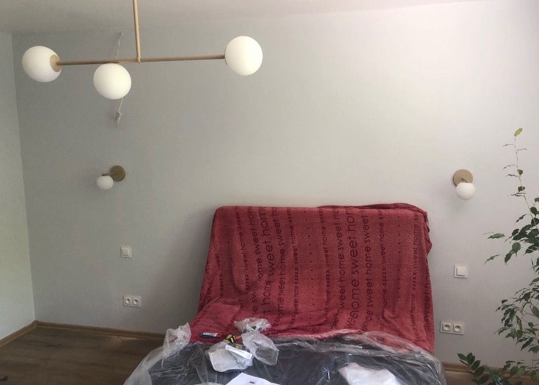 Montaż lamp: wisząca lampa sufitowa z trzema białymi kulami i dwie kinkiety ścienne z białymi kulami, w pokoju z zabezpieczonym meblem folią, widoczne gniazdka elektryczne.