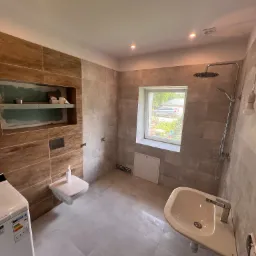 Nowoczesna łazienka w trakcie wykończenia: płytki imitujące drewno, wisząca toaleta, umywalka, pralka i półka wnękowa w ścianie z narzędziami.